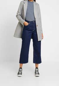 Cappotto di lana grigio sopra un maglione di maglia blu a trama, abbinato a jeans a gamba larga blu scuro e sneakers in bianco e nero.