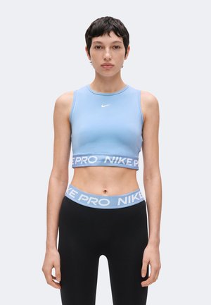 Młoda kobieta o krótkich ciemnych włosach nosi jasnoniebieski krótki top Nike i czarne legginsy z niebieskim paskiem Nike Pro, stoi na jednolitym tle.