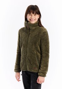 Usmívající se mladá dívka v olivově zelené huňaté bundě s vysokým límcem a černých kalhotách s hvězdným vzorem, stojící na bílém pozadí.