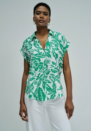 Femme portant une chemise blanche à manches courtes avec un imprimé floral vert et un pantalon blanc, debout devant un fond clair uni.