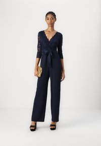 Un mono de encaje azul marino con mangas tres cuartos, escote en V, cintura ajustada y piernas anchas. Combinado con un bolso dorado y sandalias de plataforma negras.