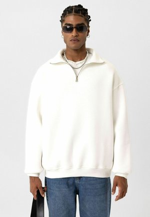 Witte ritssluiting sweatshirt met een hoge kraag, zachte textuur en een relaxte pasvorm, gecombineerd met blauwe jeans en gelaagde accessoires.