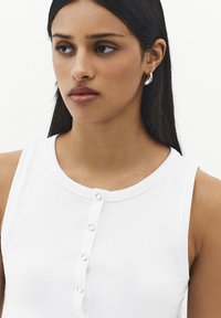 Joven mujer con cabello lacio oscuro lleva una blusa blanca sin mangas con botones y pendientes de aro plateados, mirando ligeramente hacia un lado.
