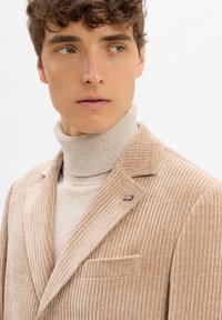 Beige Corduroy-Blazer mit geripptem Textur, Reverskragen und Tasche; kombiniert mit einem hellbeigen Rollkragenpullover. Nadel-Detail am Revers.