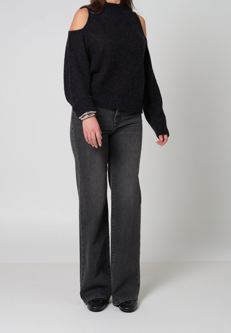 Donker marineblauwe gebreide trui met een cold shoulder ontwerp, gecombineerd met hoog-geprijsde, flared zwarte jeans en zwarte instapschoenen. Zilveren armbanden om de pols.