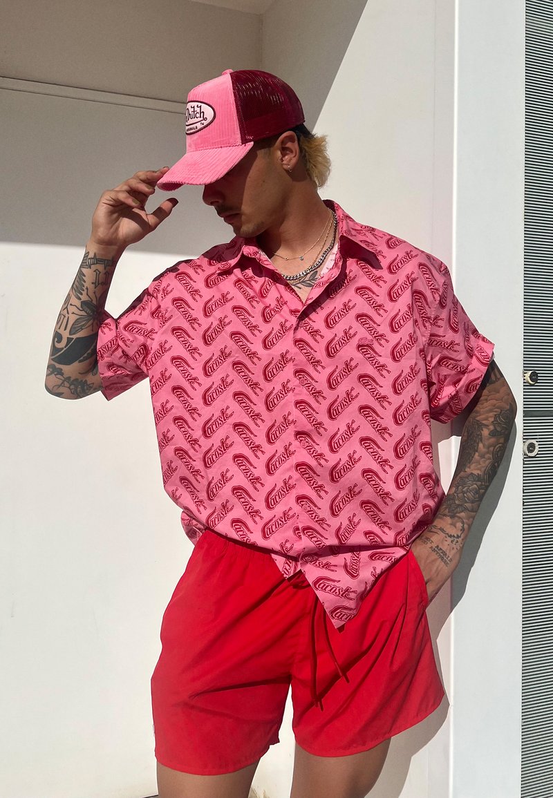 Man met tatoeages die een roze gestreept shirt, rode shorts en een roze en rode pet draagt, poseert terwijl hij met één hand de pet aanpast.
