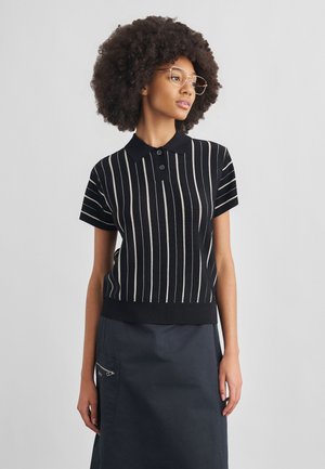 Νεαρή γυναίκα με κούρεμα αφρο, που φοράει διάφανα γυαλιά, μαύρο πόλο με κοντά μανίκια και λευκές κάθετες ρίγες, καθώς και σκουρόχρωμη φούστα με τσέπη με φερμουάρ.