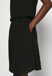 Schwarzes kurzes Kleid aus weichem Stoff, mit einem elastischen Bund und seitlichen Taschen. Der Saum ist leicht abgerundet für eine entspannte Passform.