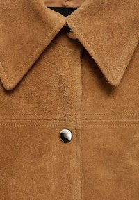 Bruine suède jas met een puntige kraag, een zilveren drukknopsluiting en zichtbare stikseldetails aan de voorkant. Zachte textuur.