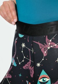 Zwarte legging met een glanzende tailleband, die kleurrijke hemelse patronen vertoont, waaronder vlinders en geometrische vormen in turkoois en paars.