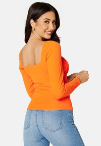 Oranje geribde lange mouw top met een vierkante halslijn, met geplooide details op de schouders, gecombineerd met lichtblauwe jeans.