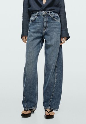 Person trägt hoch taillierte, weit geschnittene blaue Jeans mit vorderen Taschen und schwarzen offenen Sandalen mit Absatz, steht vor einem einfarbigen Hintergrund.