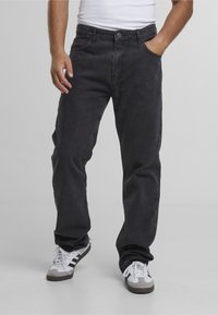 Homme portant un jean noir ample, un débardeur blanc, et des baskets blanches à rayures noires, debout, une main dans la poche.