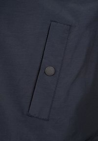 Marine jakke med en struktureret overflade, der har en slank lomme med en rund mærke-knapdetalje. Enkel, funktionel design.