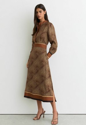 Kobieta ubrana w sukienkę midi z długim rękawem, z brązowym i beżowym nadrukiem przypominającym skórę gadów, stoi na tle białej ściany, ma na sobie beżowe sandały na wysokim obcasie.