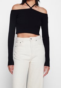 Top corto nero a costine con spalle scoperte e maniche lunghe, abbinato a pantaloni larghi a vita alta di colore bianco sporco.