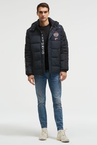 Jonge man staand, gekleed in een donkerblauw gewatteerd jack met patches, een zwart shirt, blauwe spijkerbroek en beige sneakers, op een witte achtergrond.