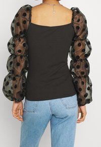 Top nero aderente con scollo quadrato e maniche trasparenti a palloncino con pois neri, abbinato a jeans azzurri chiari.