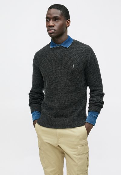 Temno sivi pleteni pulover z rebrastimi konci, nošen čez svetlo modro jeans srajco. Svetlo obarvane hlače s žeparji dopolnjujejo obleko.