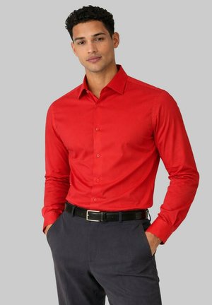 Homme portant une chemise rouge à manches longues ajustée rentrée dans un pantalon gris foncé avec une ceinture noire, les mains dans les poches, fond neutre.