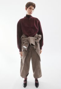 Blusa bordeaux con colletto alto abbinata a pantaloni a gamba larga marroni e testurizzati, cinte in vita. Scarpe in pelle nere.