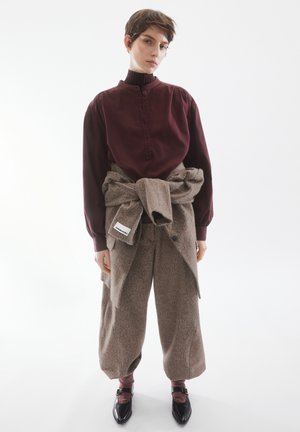 Blusa bordeaux con colletto alto abbinata a pantaloni a gamba larga marroni e testurizzati, cinte in vita. Scarpe in pelle nere.