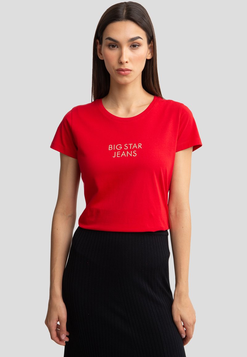 Raudona medvilninė marškinė su trumpomis rankovėmis ir apvaliu kaklu. Priekyje yra balta "BIG STAR JEANS" tekstas, derinamas su juoda raukšlėta sijonu.