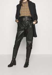 Schwarze Leder-Hosenträger mit hoher Taille und weitem Schnitt, kombiniert mit einer blumenmusternden Bluse und einem schwarzen Blazer. Knielange schwarze Stiefel mit Absatz.