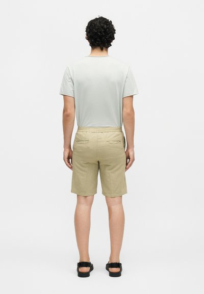 Άνδρας με σγουρά μαλλιά που φορά ανοιχτό γκρι μπλουζάκι, μπεζ σορτς με πίσω τσέπες και μαύρα σανδάλια, στέκεται γυρισμένος με την πλάτη σε λευκό φόντο.