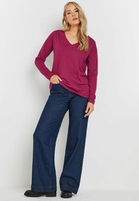 Langärmliges, V-Ausschnitt-Oberteil in tiefem Magenta, kombiniert mit weit geschnittenen, dunkelblauen Jeans. Hat eine lockere Passform und eine glatte Textur.