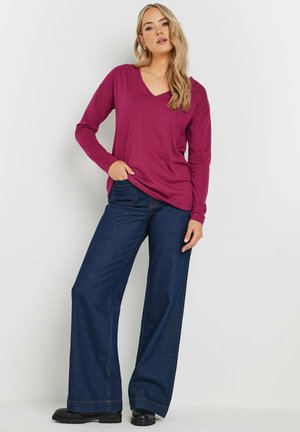 Maglietta a maniche lunghe e scollo a V in un profondo magenta, abbinata a jeans scuri a gamba larga. Presenta una vestibilità morbida e una consistenza liscia.