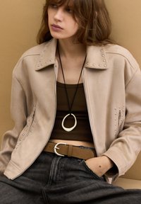 Giacca in suede beige con borchie argento sul colletto e sui lati; indossata sopra un top marrone, abbinata a jeans scuri e una cintura marrone.