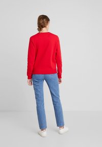 Sudadera roja de manga larga, con puños y dobladillo acanalados, combinada con vaqueros azules. El modelo está de espaldas, mostrando la parte trasera del atuendo.