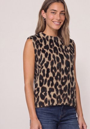 Kvinde smilende iført ærmeløs, lodden leopardprint top og blå jeans, stående foran en ensfarvet baggrund.