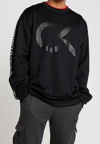 Sweat-shirt noir avec un grand logo CK en relief, manches longues, et le texte "Calvin Klein" sur la manche. Associé à un pantalon cargo gris.