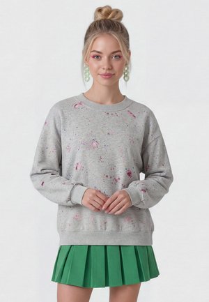 Ung kvinde med blondt hår i en knold bærer grønne kædeøreringe, en grå sweatshirt med pinke malerpletter og et grønt plisseret skørt.