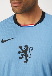 Maglia Nike azzurro chiaro con tessuto testurizzato. Presenta un emblema di leone nero e un collo a girocollo nero accentuato da strisce rosse e blu.