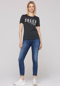Camiseta de manga corta gris oscuro con texto blanco, combinada con jeans azules ajustados y zapatillas blancas, mostrando un atuendo casual.