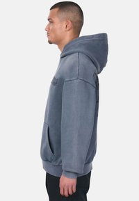 Helle graue Kapuzenjacke mit einer Fronttasche, verstellbarem Zugband an der Kapuze und gerippten Bündchen. Sichtbares Logo auf der Brust und glattes Gewebe.