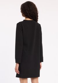 Woman with curly hair wears a simple black long-sleeve dress, shown from the back against a white background.