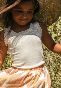 Witte geribde tanktop met franje, gecombineerd met een multikleurige rok met abstracte patronen in oranje, geel en wit. Grote strooien hoed.