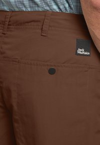 Bruine outdoorbroek van duurzaam materiaal, met een achterzak met een zwarte drukknoop en een Jack Wolfskin-logo label.