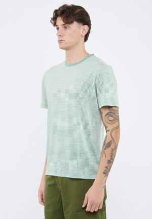 Giovane uomo in piedi, che indossa una maglietta a righe verde menta e bianca e pantaloni verde oliva, con tatuaggi visibili sul braccio destro.