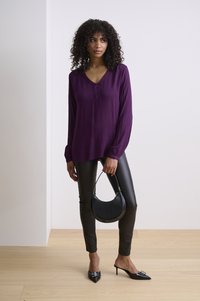 Lila Langarmbluse mit V-Ausschnitt, schwarze Lederimitat-Hose und spitze schwarze Absätze. Eine runde schwarze Handtasche rundet das Outfit ab.