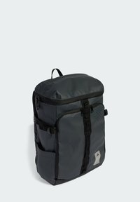 Sac à dos en nylon noir avec une fermeture roulée, une poche zippée à l'avant, des poches en mesh sur les côtés et un détail de design texturé au-dessus à l'avant.