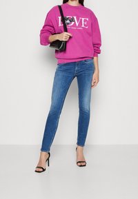 Sudadera fucsia con estampado de "LOVE", combinada con unos jeans ajustados azules. La modelo lleva tacones negros con tiras y lleva un pequeño bolso negro.