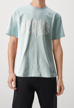 Vaaleansininen puuvillainen t-paita, jossa on rohkea valkoinen graafinen teksti "pequs" tyylitellyllä fontilla. Rentoa istuvuutta, lyhyet hihat ja pyöreä pääntie.