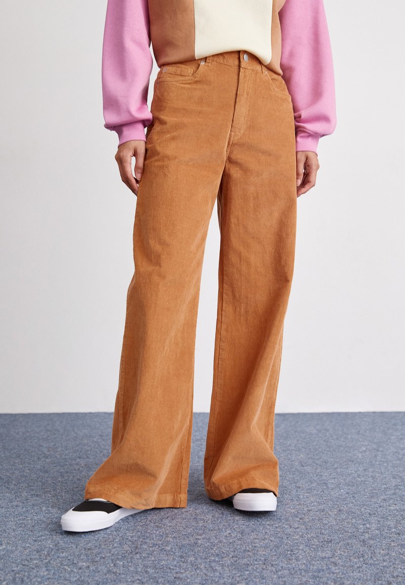 Pantalons à larges jambes en velours côtelé couleur marron chaud, avec une finition texturée, taille haute et poches avant, associés à des baskets blanches.