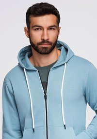 Jasnoniebieska, zapinana na zamek bluza z kapturem z białym sznurkiem, z przednim zamkiem i dwiema dolnymi kieszeniami. Noszona na ciemnozieloną koszulę.