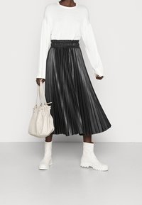 Svart plisserad läderskjorta med elastisk midja, ihopparad med en vit långärmad topp. Vita ankelskortsstövlar och en beige handväska ger kontrast.